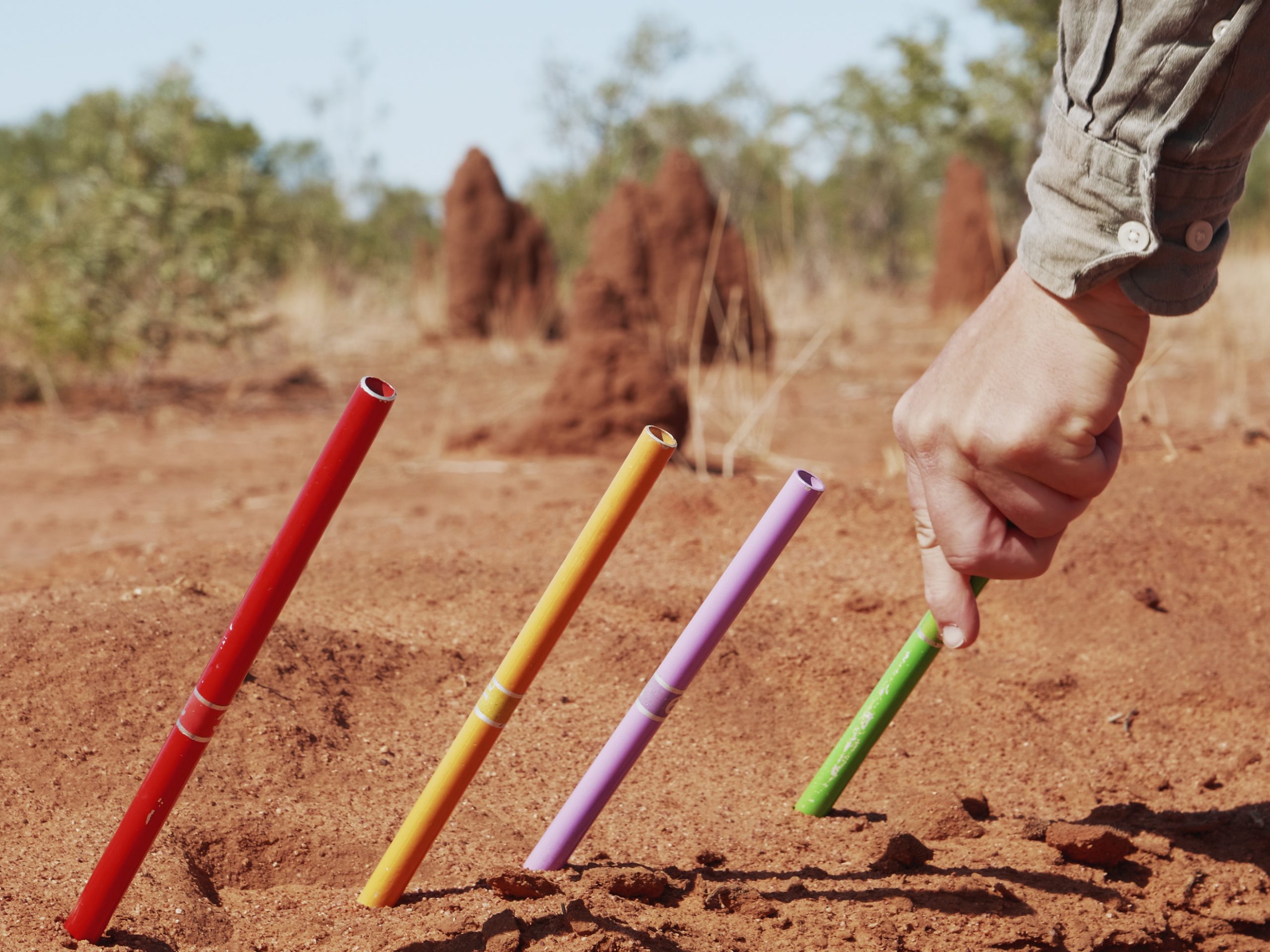 a white hand inserts a measurement tool into red earth. The tools are red, yellow, lilac and green.