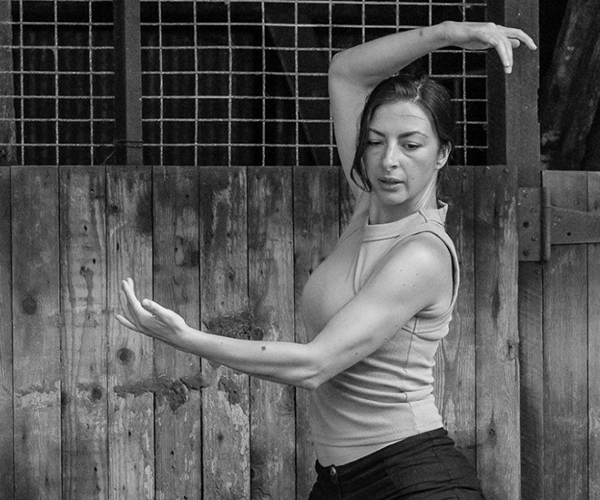 A black and white photo of Gulsen Ozer in a dance pose, with one arm curved overhead and the other scooped upward in front of her.