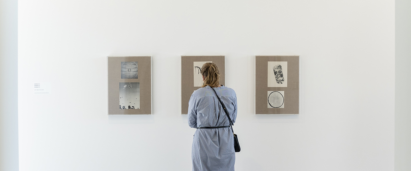 A museum visitor observes artwork at the David Noonan exhibition. Before them are collage on paper artworks hung framed on a white gallery wall. The visitor wears a dress with a pattern of thin blue and white stripes.